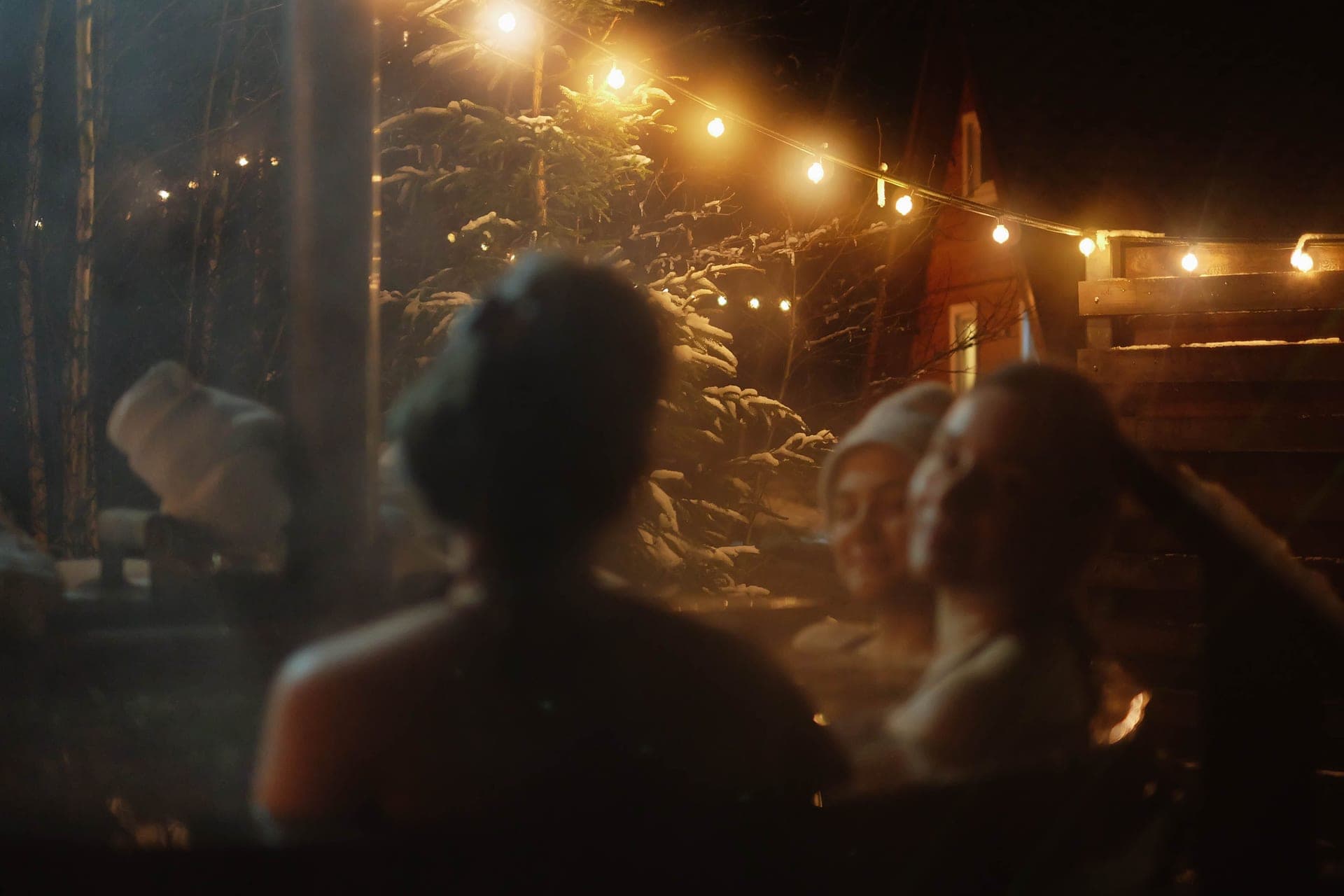 People in an outdoor hot tub at night surrounded by snow and string lights